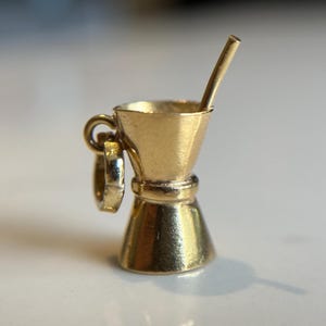 May include: A gold-colored charm in the shape of a coffee maker, featuring a handle, a spout, and a small rod inside. The charm has a polished finish and is likely made of metal.