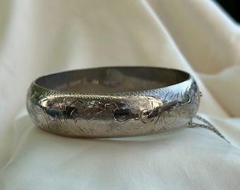 May include: A silver bangle bracelet with an intricate floral design. The bracelet is smooth and shiny, and the floral pattern is etched into the metal.
