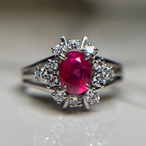 May include: A silver ring with an oval ruby centre stone, encircled by a cluster of smaller diamonds. The ring has a split shank design. The gemstones are set against a neutral background, creating a striking contrast.