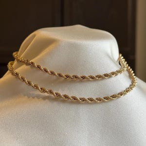 May include: Two gold-coloured rope chain necklaces displayed on a white satin surface. The necklaces are made of twisted metal links, creating a textured, rope-like appearance. The chains are close together, showcasing the design.