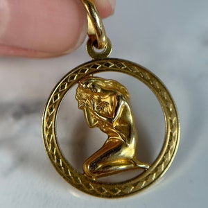 May include: A gold pendant featuring a detailed, kneeling woman figure with flowing hair, set within a circular frame with a diamond pattern.