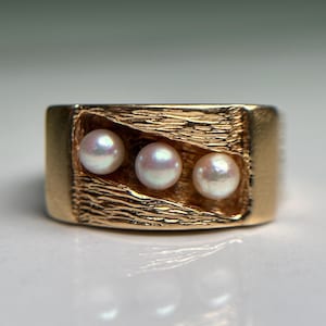 May include: A gold-colored ring featuring three small, round pearls set in a textured, rectangular design. The ring has a thick band and a polished finish, reflecting light.
