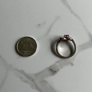 以下が含まれることがあります： ピンクの石と小さな透明な石がついたシルバーリング。リングはサイズ比較のためにカナダの5セント硬貨の隣に置かれています。