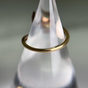 May include: A close-up shot of a gold-colored ring. The ring is thin and appears to be made of metal. The ring is displayed on a clear, cone-shaped stand, with a blurred background.