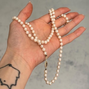 May include: A pearl necklace with a gold clasp. The necklace features numerous small, round, creamy white pearls. The clasp is gold-coloured with an oval design. The necklace is displayed on an open hand.