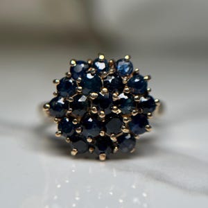 May include: A gold ring with a cluster of dark blue gemstones. The gemstones are closely set, creating a rounded, flower-like design. The ring's band is plain, polished gold.