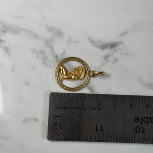 May include: Gold pendant charm featuring a woman lying down inside a circle with a braided border. The pendant is approximately 2 cm in diameter.