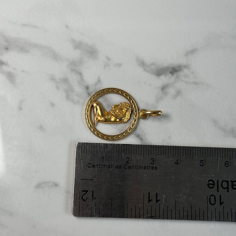 May include: Gold pendant charm featuring a woman lying down inside a circle with a braided border. The pendant is approximately 2 cm in diameter.