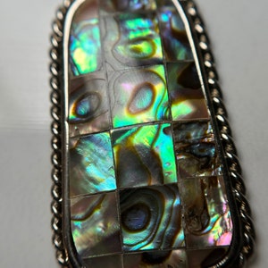 May include: A silver pendant with a rectangular abalone shell inlay. The shell is iridescent with shades of green, blue, and purple.