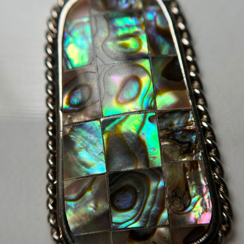 May include: A silver pendant with a rectangular abalone shell inlay. The shell is iridescent with shades of green, blue, and purple.
