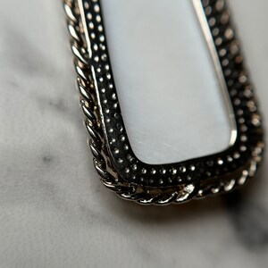 May include: A silver-toned rectangular frame with a white center and a rope-like border.