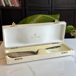 May include: An open, cream-colored box with a silver cake server inside. The server has an ornate handle and a wide, flat blade. The box is lined with a pale yellow fabric and has gold-colored clasps. The text "DAVID-ANDERSEN" is visible inside the box.