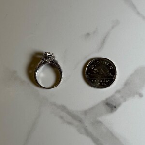 May include: A silver engagement ring with a large center diamond and smaller diamonds set in the band. The ring is next to a Canadian 2012 Toonie coin for size comparison.