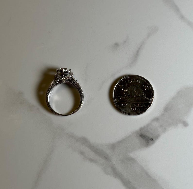 May include: A silver engagement ring with a large center diamond and smaller diamonds set in the band. The ring is next to a Canadian 2012 Toonie coin for size comparison.