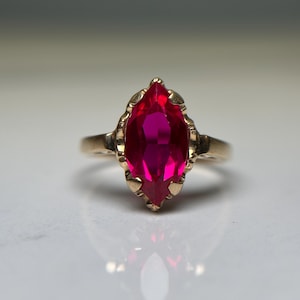 May include: A gold ring featuring a central marquise-cut vibrant pinkish-red gemstone held in a decorative prong setting. The band is smooth and metallic, showcasing a polished finish that complements the faceted stone's elongated shape.