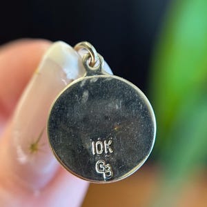Puede incluir: Un colgante circular de color dorado con un peque&ntilde;o lazo en la parte superior. El colgante tiene las marcas "10K" y "G3" estampadas en su superficie. El fondo est&aacute; borroso, con una mano sosteniendo el colgante.