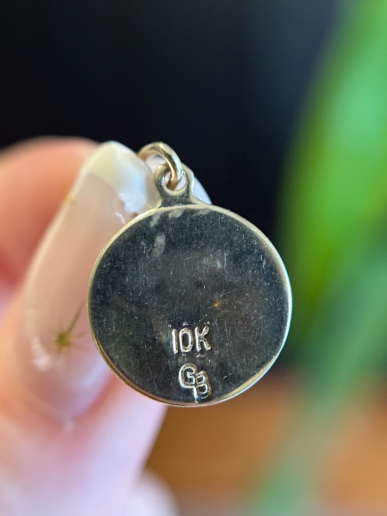 Puede incluir: Un colgante circular de color dorado con un peque&ntilde;o lazo en la parte superior. El colgante tiene las marcas "10K" y "G3" estampadas en su superficie. El fondo est&aacute; borroso, con una mano sosteniendo el colgante.