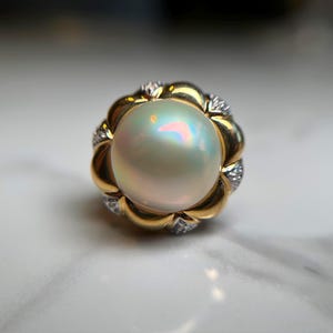 May include: A gold ring featuring a large, iridescent pearl. The pearl is surrounded by a gold setting with a floral design, accented with small, sparkling gemstones. The ring's design is elegant and eye-catching.
