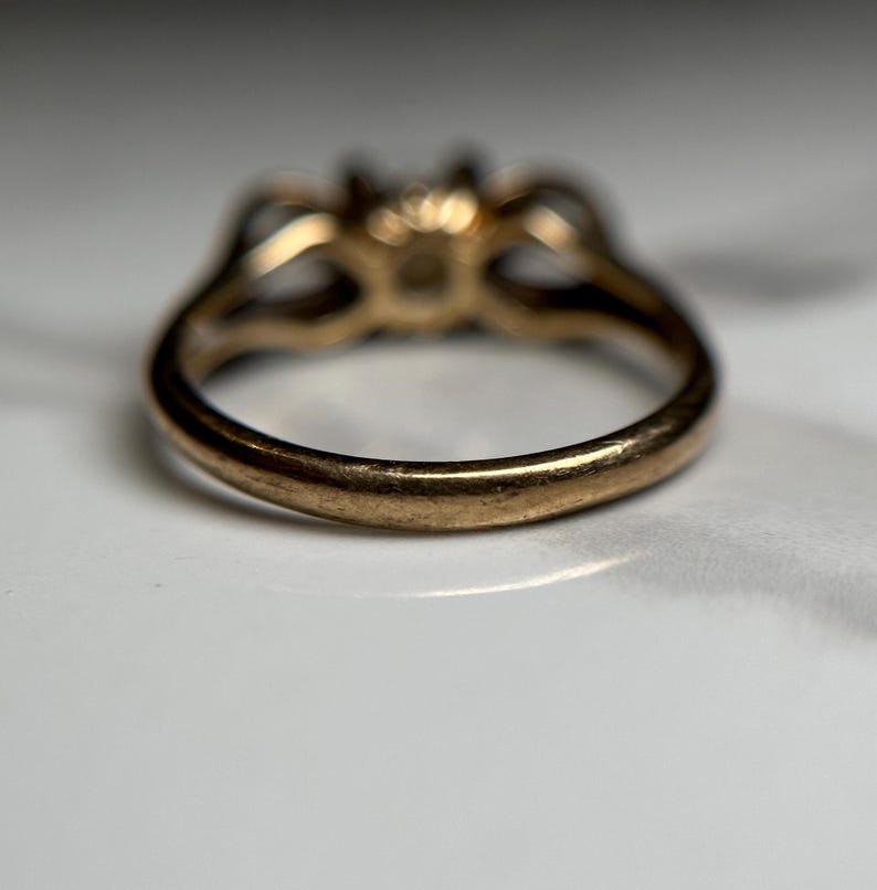 Puede incluir: Un anillo de oro con engaste de tres piedras. El anillo se muestra desde abajo, mostrando la banda lisa y redondeada.