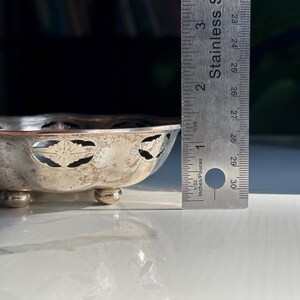 May include: A silver bowl with a scalloped edge and cut-out floral design. The bowl is sitting on three small feet and is approximately 5 inches in diameter.