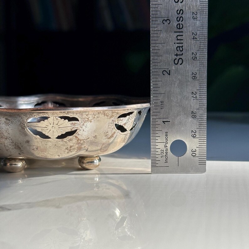 May include: A silver bowl with a scalloped edge and cut-out floral design. The bowl is sitting on three small feet and is approximately 5 inches in diameter.