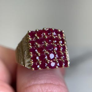 May include: A gold ring featuring a square-shaped cluster of twenty-five round, deep red rubies set in a grid pattern. The band of the ring has a textured, ribbed finish on the sides, providing a contrast to the smooth, faceted surfaces of the stones.