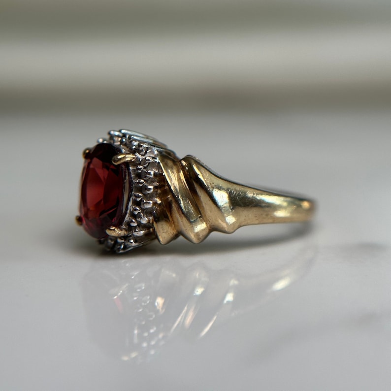 May include: A gold ring featuring a large, oval-shaped, red gemstone surrounded by smaller, white gemstones.