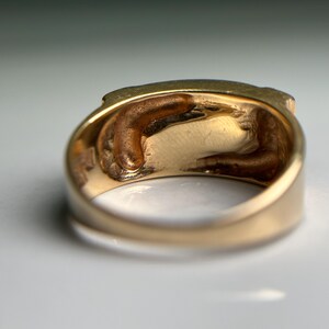 May include: A close-up of a gold ring with a detailed interior design. The ring has a thick band and a textured interior, with a slightly worn appearance. The gold color is warm and lustrous, suggesting a vintage or antique piece.