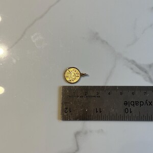 Puede incluir: Un peque&ntilde;o colgante redondo de oro con un dise&ntilde;o ornamentado, de aproximadamente 1,3 cm de di&aacute;metro. El colgante tiene un peque&ntilde;o lazo para una cadena y se muestra junto a una regla para la escala. El fondo es una superficie de m&aacute;rmol blanco.