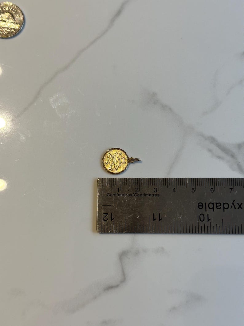 Puede incluir: Un peque&ntilde;o colgante redondo de oro con un dise&ntilde;o ornamentado, de aproximadamente 1,3 cm de di&aacute;metro. El colgante tiene un peque&ntilde;o lazo para una cadena y se muestra junto a una regla para la escala. El fondo es una superficie de m&aacute;rmol blanco.
