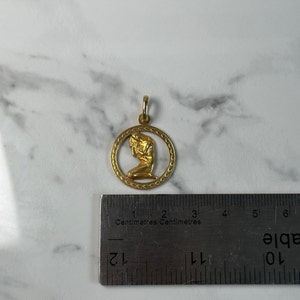 May include: Gold pendant featuring a woman kneeling in a circle with a decorative border. The pendant is 2 cm in diameter.
