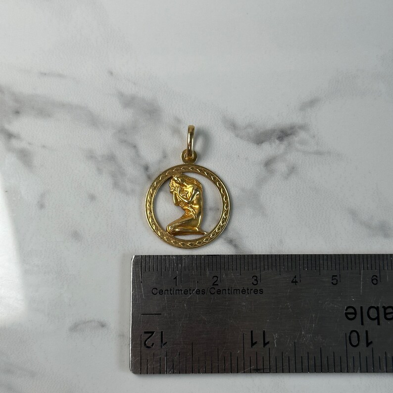 May include: Gold pendant featuring a woman kneeling in a circle with a decorative border. The pendant is 2 cm in diameter.