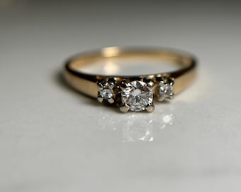 May include: A gold ring with three clear diamonds. The center diamond is larger than the two side diamonds. The ring is set on a white surface.