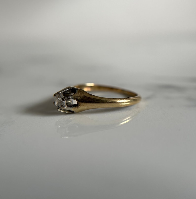 May include: A gold ring with a single, clear, square-shaped gemstone set in a prong setting.