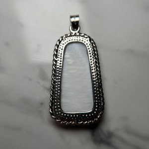 May include: A silver pendant with a white mother of pearl inlay. The pendant has a rope-like border and a textured surface.