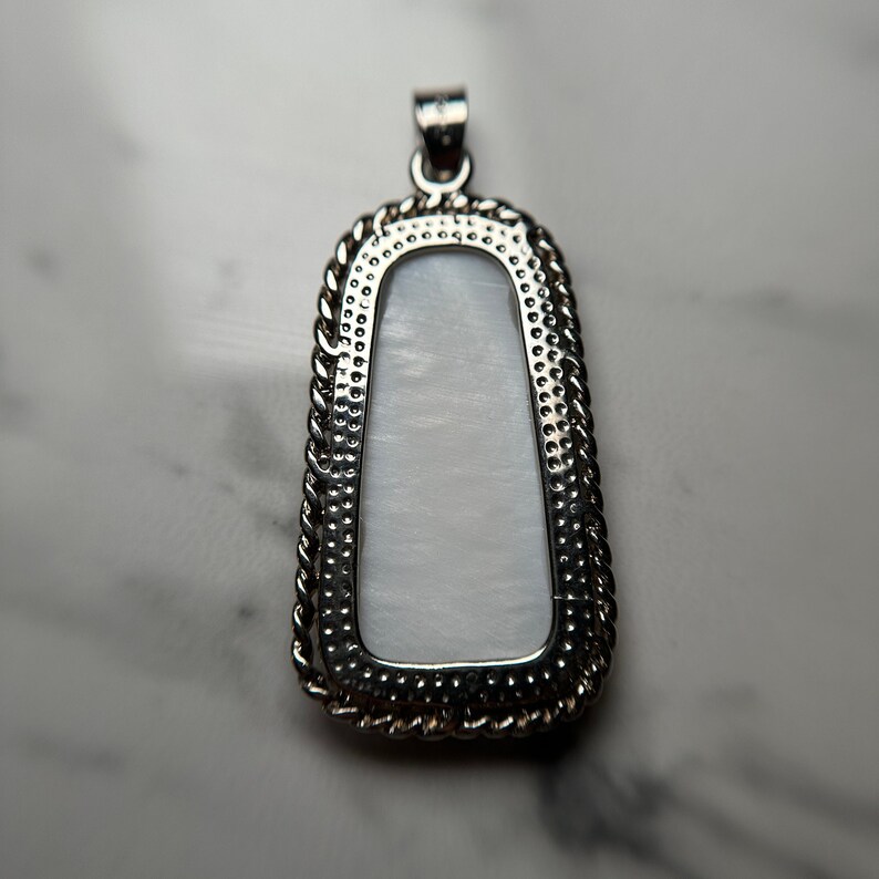 May include: A silver pendant with a white mother of pearl inlay. The pendant has a rope-like border and a textured surface.