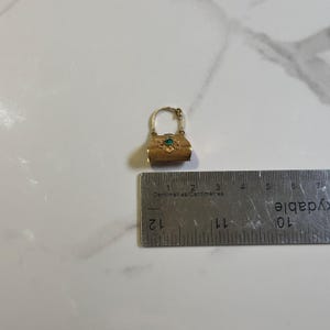 May include: A small, gold-colored purse charm with a handle and a decorative floral design. The charm features a small, green gemstone in the center. A ruler is placed next to the charm, showing measurements in centimeters.