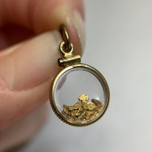 May include: A gold-colored pendant with a clear, circular center containing several small gold nuggets. The pendant has a loop at the top for a chain and a decorative, textured edge.