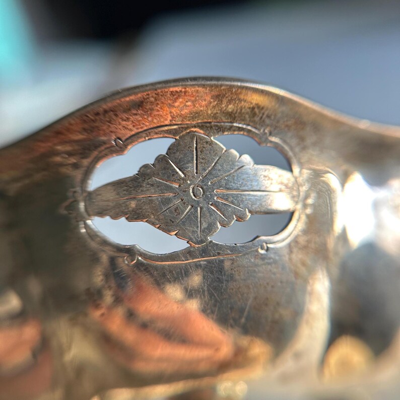 May include: Close-up of a silver spoon with a decorative cutout design featuring a stylized flower with a circle in the center.