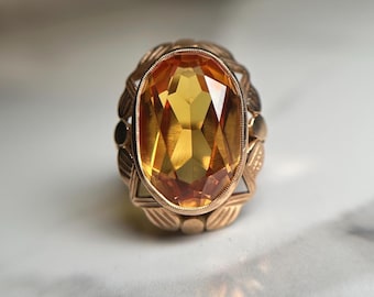 May include: A gold ring featuring a large, oval-cut, amber-colored gemstone set in a decorative metal frame. The band and setting display a textured, leaf-like pattern surrounding the central stone, which exhibits multiple facets and a warm, golden hue.