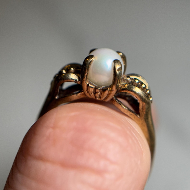 Puede incluir: Un anillo de oro con una perla blanca en el centro. El anillo tiene un dise&ntilde;o delicado e intrincado.
