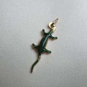 May include: A gold-colored lizard charm with green enamel detailing. The lizard is depicted in a stylized pose, with its legs outstretched and tail curved. The charm has a small loop for attaching to a necklace or bracelet.