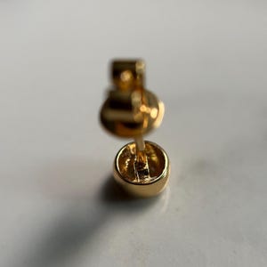 May include: Close-up of a gold stud earring. The earring has a small, round, polished design. The post and backing are also gold. The earring is set against a white background.