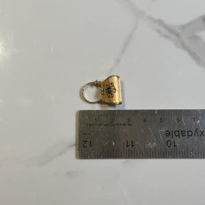 May include: A gold-colored earring with a textured surface and a small green gemstone in the center. The earring is approximately 1 inch in length and is next to a ruler.