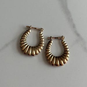 May include: A pair of gold-colored hoop earrings. Each earring has a textured, ridged design along the outer curve. The earrings are displayed on a white and gray marble surface. The earrings are a classic jewelry item.