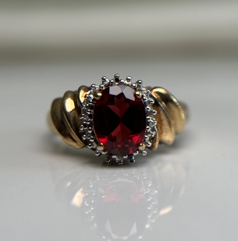 May include: A gold ring featuring a large, oval-shaped, red gemstone surrounded by smaller, white gemstones.