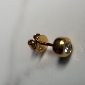 May include: A single gold earring with a round, bezel-set diamond. The earring post and backing are also gold. The backing is stamped with the number "750". The earring is on a white surface.