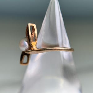 May include: A gold-toned ring featuring a pearl and a decorative design. The ring's design includes a curved band and an openwork element, with a small, round pearl. The ring is displayed on a clear, conical stand.