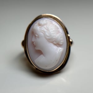 May include: A gold ring with a cameo of a woman's profile carved in white shell. The cameo is set in a gold oval frame.