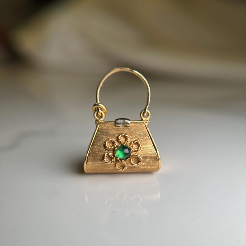 May include: A small, gold-colored handbag charm with a textured surface. The charm features a flower design with a green gemstone center. The handle is a curved, gold-colored wire.